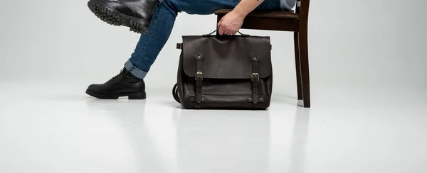 El hombre en pantalones vaqueros azules y botas negras se sienta en una silla con un bolso de cuero marrón para hombre para documentos y un portátil en un piso blanco. Bolso de cuero para hombre, bolsos de mensajero, maletín hecho a mano. — Foto de Stock