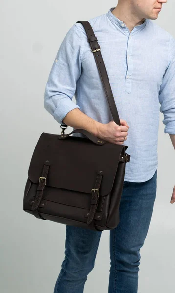 Brązowa męska torba na ramię na dokumenty i laptop na ramionach mężczyzny w niebieskiej koszuli i dżinsach z białym tłem. Torebka męska skórzana, ręcznie robiona walizka. — Zdjęcie stockowe