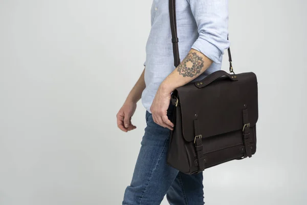 Brązowa męska torba na ramię na dokumenty i laptop na ramionach mężczyzny w niebieskiej koszuli i dżinsach z białym tłem. Torebka męska skórzana, ręcznie robiona walizka. — Zdjęcie stockowe