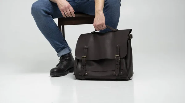 El hombre en pantalones vaqueros azules y botas negras se sienta en una silla con un bolso de cuero marrón para hombre para documentos y un portátil en un piso blanco. Bolso de cuero para hombre, bolsos de mensajero, maletín hecho a mano. — Foto de Stock