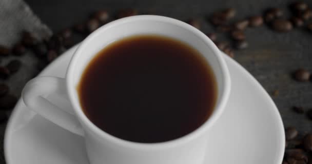 Fehér csésze kávé pörkölt kávébabbal szétszórva egy fa asztalon. Egy bögre friss fekete kávé. Friss arab pörkölt kávébab. Remek kezdet a tökéletes reggelhez. Eszpresszó, americano, doppio. — Stock videók