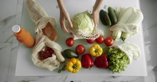 La mujer saca la col de la bolsa de comestibles reutilizable con verduras en una mesa con pimientos, tomates y pepinos en la cocina. Residuos cero, plástico gratis, compras de comestibles. Comprador de algodón de malla. — Vídeos de Stock