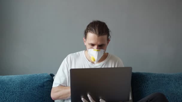 Hombre en camiseta y máscara médica, respirador está trabajando de forma remota en casa en un sofá con el ordenador portátil hablando con amigos o socios a través de la webcam. pandemia de Coronavirus. Quédate en casa, COVID-19, cuarentena.. — Vídeos de Stock