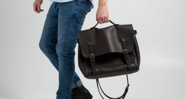 Kahverengi erkek omuz çantası. Mavi gömlekli ve beyaz arka planlı kot pantolonlu bir adam tarafından tutulan belgeler ve dizüstü bilgisayar için. Çanta, erkek derisi el yapımı çanta..