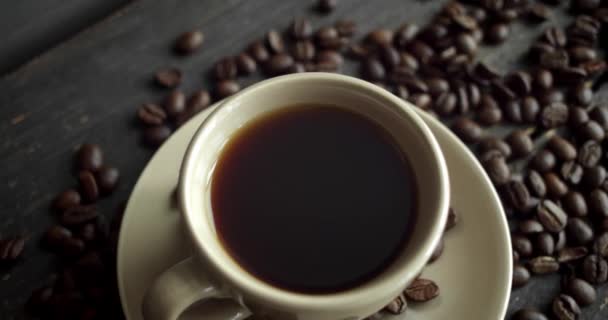 Καφές με καβουρδισμένους κόκκους καφέ σκορπισμένους σε ξύλινο τραπέζι. Μια κούπα φρέσκου μαύρου καφέ. Φρέσκοι καβουρδισμένοι κόκκοι καφέ. Ωραία αρχή του τέλειου πρωινού. Espresso, americano, doppio. — Αρχείο Βίντεο