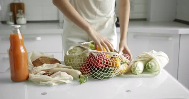 Vrouw neemt gele en rode peper uit herbruikbare boodschappentas met groenten op een tafel in de keuken thuis na boodschappen doen. Geen afval en plastic vrij concept. Mesh katoenshopper. — Stockvideo