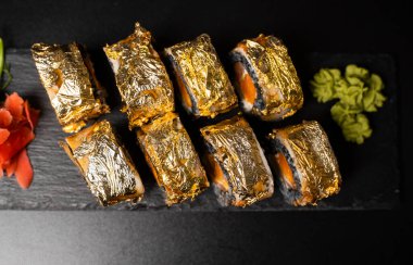 Özel suşi rulosu, mürekkep balığı mürekkebi, taze somon, krem peynir, biber kabağı, yılan balığı, unagi sosu ve kırmızı zencefilli yenilebilir altın yaprak, siyah masa ve arka plandaki kare tabakta wasabi..