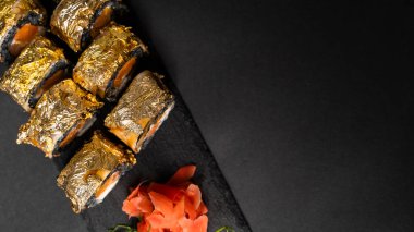 Özel suşi rulosu, mürekkep balığı mürekkebi, taze somon, krem peynir, biber kabağı, yılan balığı, unagi sosu ve kırmızı zencefilli yenilebilir altın yaprak, siyah masa ve arka plandaki kare tabakta wasabi..