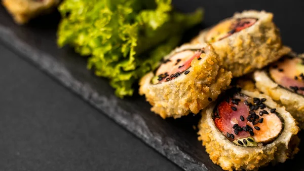 Rolo de sushi personalizado em tempura com nori, salmão fresco, atum, abacate, caviar de masago, regado com molho de abacaxi com salada derramando como decoração em uma placa preta em uma mesa preta e fundo. — Fotografia de Stock