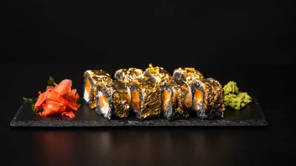 Custom sushi roll with nori, cuttlefish ink, fresh salmon, cream cheese, pepper pumpkin, eel, unagi sauce and edible gold leaf with red ginger, wasabi on a square plate on black table and background.