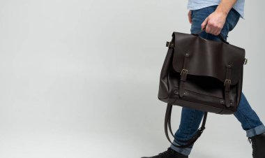 Kahverengi erkek omuz çantası. Mavi gömlekli ve beyaz arka planlı kot pantolonlu bir adam tarafından tutulan belgeler ve dizüstü bilgisayar için. Çanta, erkek derisi el yapımı çanta..