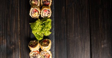 Taze somon, avokado, ananas soslu masago havyarı, ahşap masa ve arka plandaki siyah bir tabağa süsleme olarak salata serpiştirilmiş özel suşi rulosu..