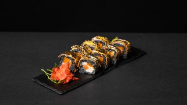 Özel suşi rulosu, mürekkep balığı mürekkebi, taze somon, krem peynir, biber kabağı, yılan balığı, unagi sosu ve kırmızı zencefilli yenilebilir altın yaprak, siyah masa ve arka plandaki kare tabakta wasabi..
