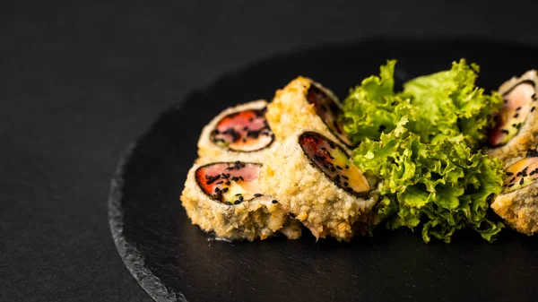 Rollo de sushi personalizado en tempura con nori, salmón fresco, atún, aguacate, caviar masago, rociado con salsa de piña con ensalada vertiendo como decoración en un plato negro sobre una mesa negra y fondo. —  Fotos de Stock