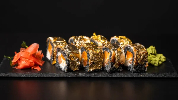 Custom sushi roll with nori, cuttlefish ink, fresh salmon, cream cheese, pepper pumpkin, eel, unagi sauce and edible gold leaf with red ginger, wasabi on a square plate on black table and background.