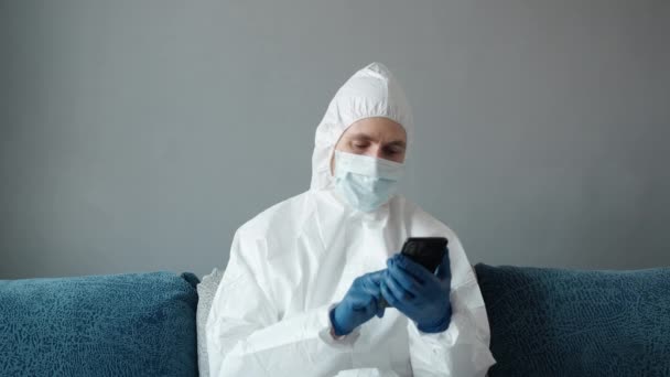 Pria berjas putih pelindung dan masker medis menggunakan telepon di rumahnya duduk di tempat tidur karena epidemi coronavirus. Pekerjaan jarak jauh selama pandemi. Tinggal di rumah selama konsep karantina COVID-19. — Stok Video