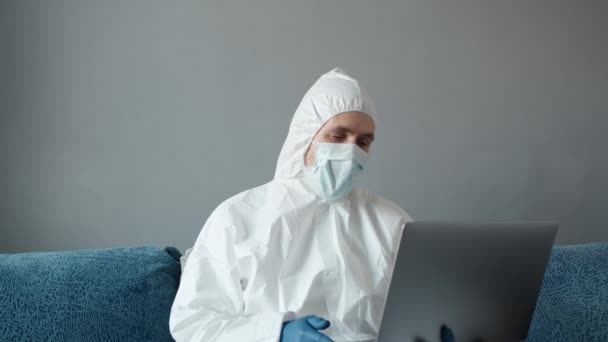 El hombre con traje blanco protector y máscara médica está trabajando desde casa en un sofá con computadora portátil debido a la pandemia de coronavirus. Trabajo remoto durante una pandemia. Permanecer en casa durante el concepto de cuarentena COVID-19. — Vídeos de Stock
