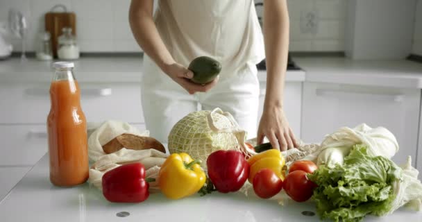 Kvinna tar fram färska grönsaker, avokado, tomater och gurka från återanvändbara matkassar med grönsaker på ett bord med paprika och sallad. Noll spill, plastfritt, dagligvaruhandel. Mesh shoppare. — Stockvideo