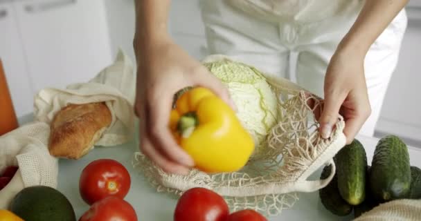 Kadın tekrar kullanılabilir bir alışveriş poşetinin içine sarı biber ve domates koydu. Sebzeli ve salatalı bir masaya. Mutfakta portakal suyu. Sıfır atık, plastiksiz, market alışverişi. Pamuk tüccarı.. — Stok video