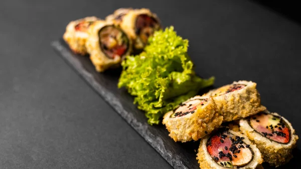 Rolo de sushi personalizado em tempura com nori, salmão fresco, atum, abacate, caviar de masago, regado com molho de abacaxi com salada derramando como decoração em uma placa preta em uma mesa preta e fundo. — Fotografia de Stock