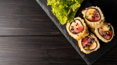 Taze somon, avokado, ananas soslu masago havyarı, ahşap masa ve arka plandaki siyah bir tabağa süsleme olarak salata serpiştirilmiş özel suşi rulosu..