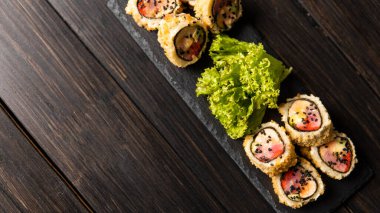 Taze somon, avokado, ananas soslu masago havyarı, ahşap masa ve arka plandaki siyah bir tabağa süsleme olarak salata serpiştirilmiş özel suşi rulosu..