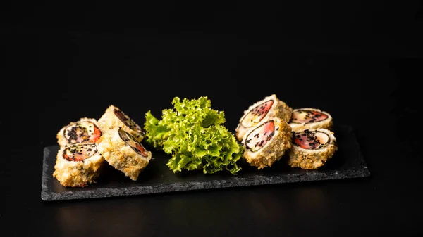 Rollo de sushi personalizado en tempura con nori, salmón fresco, atún, aguacate, caviar masago, rociado con salsa de piña con ensalada vertiendo como decoración en un plato negro sobre una mesa negra y fondo. —  Fotos de Stock