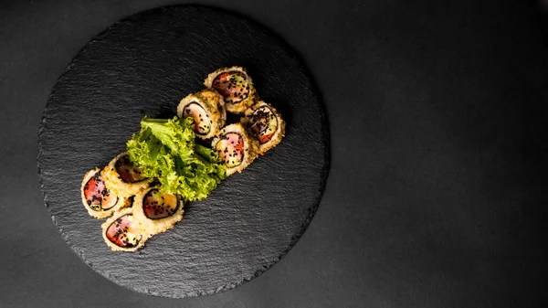 Rollo de sushi personalizado en tempura con nori, salmón fresco, atún, aguacate, caviar masago, rociado con salsa de piña con ensalada vertiendo como decoración en un plato negro sobre una mesa negra y fondo. —  Fotos de Stock