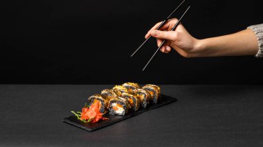 Siyah çubuklu el, mürekkep balığı mürekkebi, somon, krem peynir, biber kabağı, yılanbalığı ve zencefilli yenilebilir altın yaprak, siyah masadaki kare bir tabakta wasabi almak istiyor..