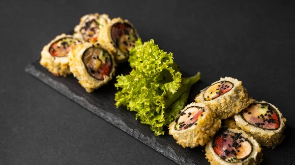 Rolo de sushi personalizado em tempura com nori, salmão fresco, atum, abacate, caviar de masago, regado com molho de abacaxi com salada derramando como decoração em uma placa preta em uma mesa preta e fundo. — Fotografia de Stock