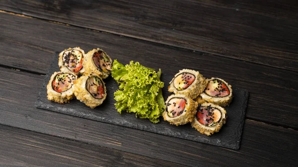 Rolo de sushi personalizado em tempura com nori, salmão fresco, atum, abacate, caviar de masago, regado com molho de abacaxi com salada derramando como decoração em uma placa preta em uma mesa de madeira e fundo. — Fotografia de Stock