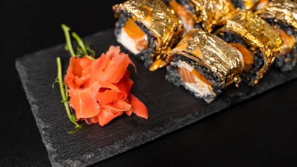 Rolo de sushi personalizado com nori, tinta de choco, salmão fresco, queijo creme, abóbora de pimenta, enguia, molho unagi e folha de ouro comestível com gengibre vermelho, wasabi em uma placa quadrada na mesa preta e fundo. — Fotografia de Stock