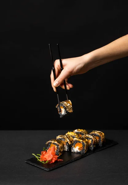 Mão com pauzinhos pretos segura um sushi. Rolo de sushi personalizado com tinta de choco, salmão, queijo creme, abóbora de pimenta, enguia e folha de ouro comestível com gengibre, wasabi em um prato quadrado na mesa preta. — Fotografia de Stock