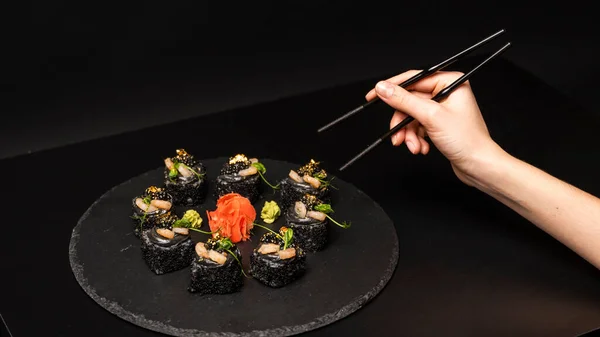 Ręka z pałeczkami chce wziąć niestandardowe sushi roll z czarnym ryżem, mięso krabowe, awokado, wędzony mus łososiowy, kawior wiosło, masago, koktajl krewetki, jadalne liście złota, imbir, wasabi na czarnym stole. — Zdjęcie stockowe