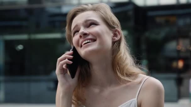 Joven mujer de negocios en una camiseta blanca es risas y hablar por teléfono con socios de negocios o amigos con un centro de negocios o centro comercial detrás. Chica feliz dice o escucha algo divertido. — Vídeos de Stock