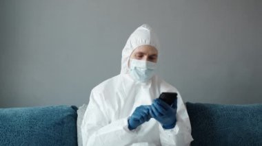 Koruyucu beyaz takım elbiseli ve tıbbi maskeli bir adam Coronavirüs salgını yüzünden evinde yatağında oturan bir telefonu kullanıyor. Salgın sırasında uzaktan çalışma. COVID-19 karantina sırasında evde kal.