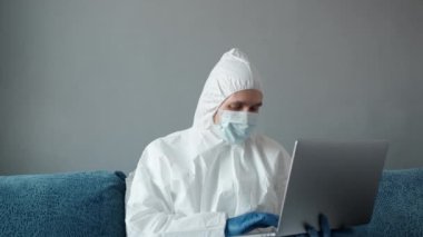 Koruyucu beyaz takım elbiseli ve tıbbi maskeli bir adam Coronavirus salgını yüzünden evinde dizüstü bilgisayarla çalışıyor. Salgın sırasında uzaktan çalışma. COVID-19 karantina sırasında evde kal.