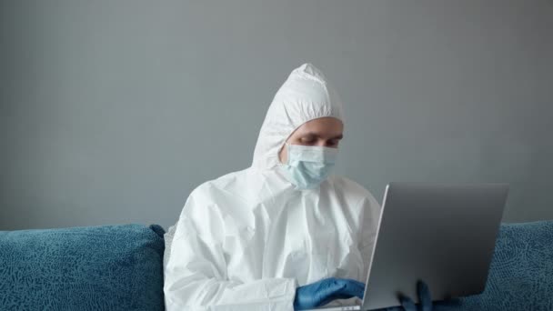 El hombre con traje blanco protector y máscara médica está trabajando desde casa en un sofá con computadora portátil debido a la pandemia de coronavirus. Trabajo remoto durante una pandemia. Permanecer en casa durante el concepto de cuarentena COVID-19. — Vídeo de stock