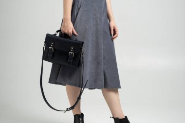 Beyaz arka planda bir kadının elindeki küçük siyah deri çanta. Omuz çantası. Beyaz gömlekli, gri etekli ve siyah çantalı bir kadın. Tarz, retro, moda, klasik ve zarafet.