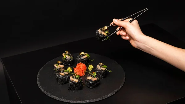 Ręka z pałeczkami trzyma sushi. Rolka sushi na zamówienie z czarnym ryżem, mięso krabowe, awokado, wędzony mus łososiowy, kawior wiosłowy, masago, koktajl krewetkowy, jadalne liście złota, imbir, wasabi na czarnym stole. — Zdjęcie stockowe