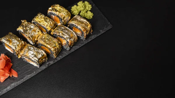 Custom sushi roll with nori, cuttlefish ink, fresh salmon, cream cheese, pepper pumpkin, eel, unagi sauce and edible gold leaf with red ginger, wasabi on a square plate on black table and background.
