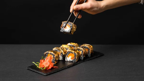 Ręka z czarnymi pałeczkami trzyma sushi. Niestandardowe sushi roll z atramentem cuttlefish, łosoś, ser śmietankowy, dynia pieprzowa, węgorz i jadalny złoty liść z imbirem, wasabi na kwadratowym talerzu na czarnym stole. — Zdjęcie stockowe