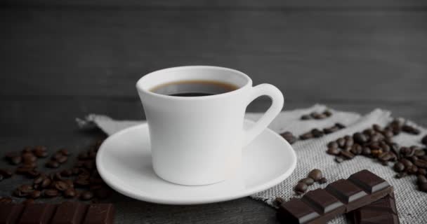 Šálek kávy s čokoládou a praženými kávovými zrny roztroušený na dřevěném stole. Černý hrnek na kávu. Čerstvé arabické pražené kávové zrno. Skvělý začátek rána. Espresso, americano, doppio.