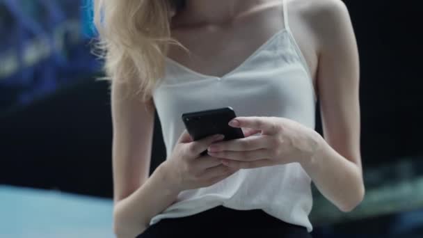 Молода ділова жінка в білій футболці використовує телефон і пише повідомлення бізнес-партнеру або друзям з бізнес-центром або торговим центром позаду. Щаслива дівчина каже або чує щось смішне . — стокове відео