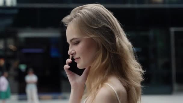 Joven mujer de negocios en una camiseta blanca es risas y hablar por teléfono con socios de negocios o amigos con un centro de negocios o centro comercial detrás. Chica feliz dice o escucha algo divertido. — Vídeos de Stock