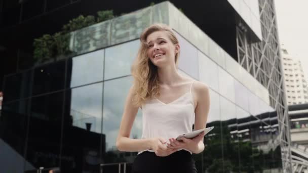 Retrato de mujer de negocios en una camiseta blanca se ríe y sostiene una tableta con un centro de negocios detrás, al aire libre. Mujer joven feliz en una reunión y esperando socio de negocios. — Vídeos de Stock
