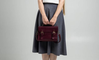 Beyaz arka planda bir kadının elinde küçük kırmızı deri bir çanta. Omuz çantası. Beyaz gömlekli, gri etekli ve kırmızı çantalı bir kadın. Tarz, retro, moda, klasik ve zarafet.