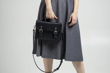 Beyaz arka planda bir kadının elindeki küçük siyah deri çanta. Omuz çantası. Beyaz gömlekli, gri etekli ve siyah çantalı bir kadın. Tarz, retro, moda, klasik ve zarafet.