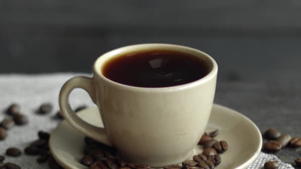 Kropla kawy upuszcza beżową filiżankę espresso z ziarnem kawy rozrzuconym na stole i płótno na drewnianym stole. Świeże pyszne kawy jest gotowy do picia. — Wideo stockowe