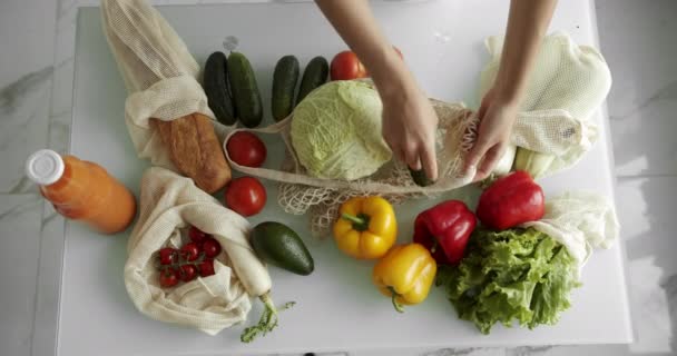 Mujer poner un pepino dentro de la bolsa de comestibles reutilizables con verduras en una mesa con pimientos, tomates y pepinos en la cocina. Residuos cero, plástico gratis, compras de comestibles. Comprador de algodón de malla. — Vídeos de Stock